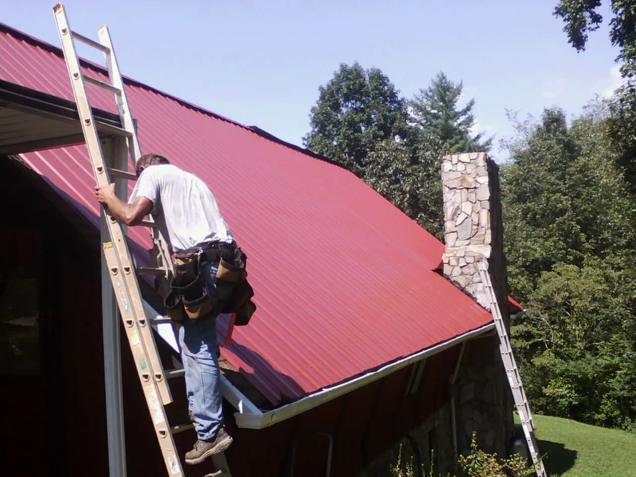 Roofer on a ladder beside a red metal roof and stone chimney — Soffit & Fascia Repair by Roofing Brothers in Redlands